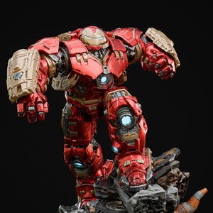 Könnte beinhalten: Eine rote und goldene Iron Man Hulkbuster-Statue. Die Figur steht auf einem felsigen Sockel mit einem Reifen und einem Verkehrskegel. Die Statue hat komplizierte Details und leuchtende blaue Akzente.
