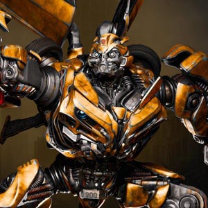 以下が含まれることがあります： 黄色と黒のロボットの詳細な画像。武器を前方に構え、複雑な機械的ディテールが特徴。背景にはぼやけた都市景観があり、戦闘シーンを暗示。武器からは光が放たれている。未来的なアクションをテーマにした画像。