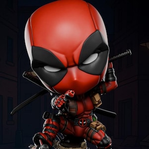 May include: A Chibi Design Deadpool figurine in a dynamic pose, clad in a red and black suit. The figure is holding two katanas and is positioned on a detailed base with the word "BOOM" visible. The figurine has a stylized, oversized head.