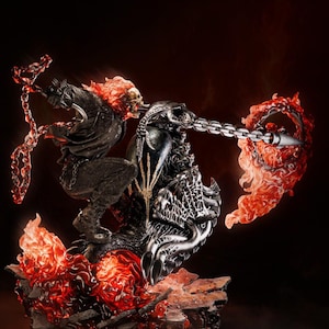May include: A detailed statue of a flaming skull figure riding a motorcycle, with a chain weapon. The figure is predominantly black and silver, with fiery red and orange accents. The base is a rocky, textured surface.