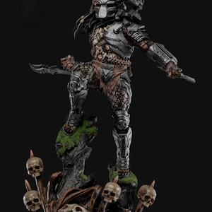 Puede incluir: Figura de acción detallada de Predator, de pie sobre una base rocosa con calaveras. La figura está vestida con una armadura metálica y lleva armas. La base presenta musgo y calaveras adicionales, creando una exhibición dinámica y amenazante.