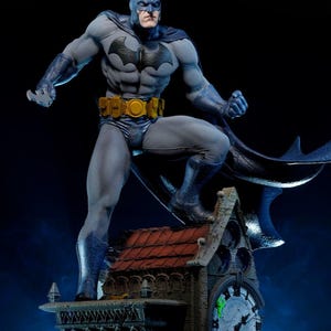 May include: A Batman statue in a dynamic pose, wearing a grey and blue suit with a black bat symbol. The figure stands on a clock tower with a red roof, a yellow utility belt, and a flowing cape. The statue is a collectible item.