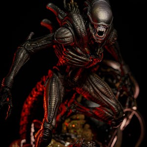 May include: A detailed, dark grey Xenomorph figure with a menacing open mouth, sharp teeth, and a textured exoskeleton. The figure is posed against a dark background with red lighting, highlighting its intricate design and alien features. A collectible item.