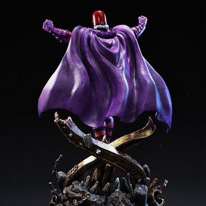 May include: A statue of a character in a purple cape and helmet, arms raised. The figure stands on a detailed base with a metallic X-shaped design. The cape is a deep purple, and the helmet is red.