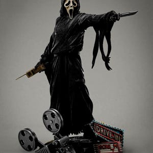 Può includere: Una figura nera con un cappuccio e una maschera, che tiene in mano un coltello e un telefono vintage. La figura è su una base con un proiettore cinematografico e un'insegna "DRIVE-IN". La maschera della figura ha un'espressione urlante.
