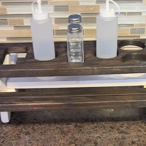 May include: A dark stained wooden picnic table-style condiment holder with two squeeze bottles, a salt and pepper shaker, and two circular cutouts. The base and legs are painted white.