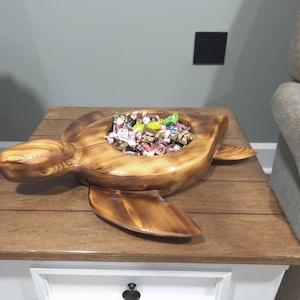 May include: A wooden turtle-shaped bowl filled with wrapped sweets. The bowl is made of light brown wood and sits on a wooden table. The turtle's head and flippers extend outward. The table is white with a brown top.