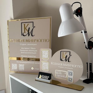Könnte beinhalten: Eine Präsentation mit Logo und Geschäftsinformationen. Sie umfasst ein rechteckiges Schild, ein rundes Schild und einen kleinen Kartenhalter, alle in Beige und Gold. Die Schilder zeigen den Firmennamen und die angebotenen Dienstleistungen.