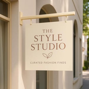 May include: A cream-coloured rectangular sign with the text "THE STYLE STUDIO" in a brown serif font, with a leaf design and the words "CURATED FASHION FINDS" below. The sign is suspended from a gold-coloured bracket.