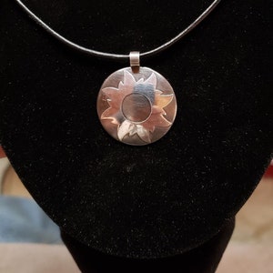 Peut inclure: Collier avec pendentif en argent. Le pendentif circulaire présente un motif de soleil. Le pendentif est suspendu à un cordon fin et foncé. Le collier est présenté sur fond noir.