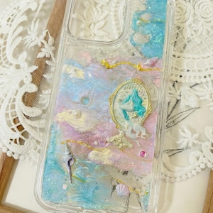 May include: Clear phone case with a mermaid design. The case has a layered design with blue, pink, and white colours, gold accents, and a mermaid illustration. Decorative elements include seashells and pearls.