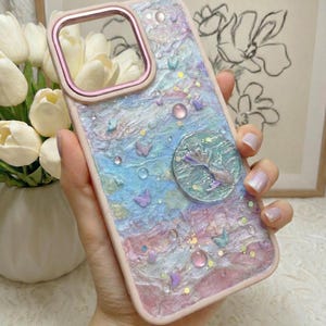 Peut inclure: Une coque de téléphone avec une surface texturée aux tons pastel mélangés de rose, bleu et violet. Le design présente de petites décorations en forme de papillon, des perles rondes et un élément circulaire représentant une queue de sirène, le tout pailleté.