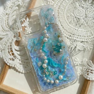 Peut inclure: Une coque de smartphone transparente avec un design texturé et marbré dans des tons bleu, violet et blanc, agrémenté de paillettes. La coque est ornée d'une fine chaîne de fausses perles blanches et de deux petites breloques en forme d'étoile.