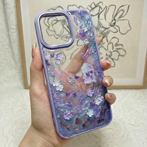 Peut inclure: Une coque de téléphone transparente avec un cadre violet, présentant un design texturé en résine violette et bleue, des petites décorations florales, des papillons miniatures et un motif de porte arquée intégré à la surface arrière.