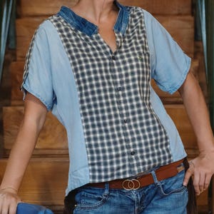 Pode incluir: Uma camisa de manga curta com gola de ganga e mangas azul claro. O painel central apresenta um padrão xadrez preto e branco. A camisa é combinada com jeans azuis e um cinto castanho com fivela dourada.