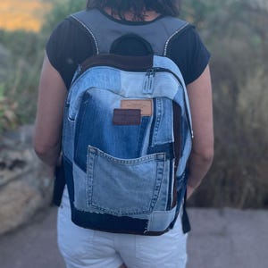 Op de afbeelding: Een denim rugzak met een patchwork ontwerp. De rugzak heeft verschillende tinten blauw denim, een bruine suède bovenkant en een voorvak. Het heeft een leren label en een ritssluiting. De bandjes zijn zwart en grijs.