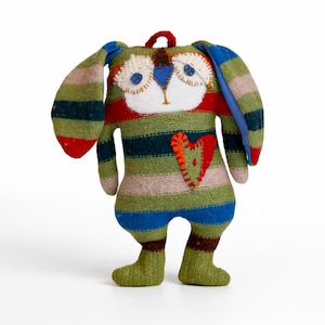 May include: A handmade soft toy creature with a striped pattern in green, blue, red, and tan. It features large ears, a heart-shaped patch, and a loop at the top. The eyes are white with blue pupils.