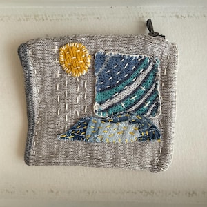 Può includere: Pochette grigia in maglia con design paesaggistico. La pochette presenta un sole giallo, un quadrato blu e verde e un paesaggio cucito blu e giallo. Con chiusura a zip.