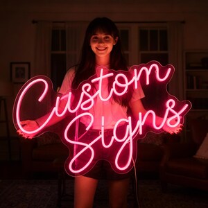May include: A pink neon sign, spelling out "Custom Signs" in a flowing script, is illuminated. The sign is held by a person. The warm lighting of the room creates a striking contrast with the neon.