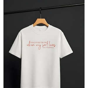 Könnte beinhalten: Weißes T-Shirt mit rotem Text "I HAVE FOUND THE ONE whom my soul loves" und "SONG OF SOLOMON 3:4". Das Shirt hängt an einem Holzbügel vor dunklem Hintergrund.