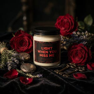 May include: A cream-colored candle in a glass jar with a black lid, surrounded by red roses and antique keys. The label reads "Light When You Miss Me" in red text. The candle is set on a dark velvet surface.