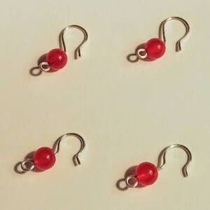 May include: Four small, handmade silver-tone metal stitch markers, each featuring a vibrant red bead. The markers have a unique hook design, perfect for marking stitches in knitting or crochet projects. The beads are a bright, translucent red.