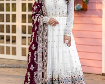 May include: A white embroidered gown with long sleeves and a flowing skirt. The gown features intricate gold and silver embellishments along the neckline, waist, and hem. A burgundy shawl with ornate detailing drapes over the shoulders. The outfit is suitable for special occasions.