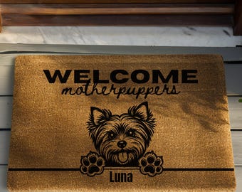 May include: A rectangular doormat made of natural fibers. The mat features the words "WELCOME motherpuppers" in black, with a graphic of a dog's face and paws. The name "Luna" is printed below the dog.