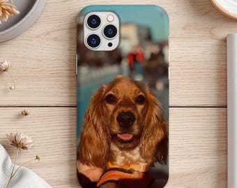 May include: A phone case featuring a golden Cocker Spaniel with a blurred background. The case has a blue sky and cityscape background. The dog is wearing an orange and black harness. The phone case is on a light wood surface.