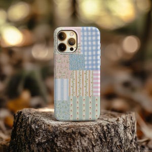 May include: A patchwork-style phone case featuring a variety of patterns, including floral prints, stripes, and gingham checks in pastel colors. The case has a camera cutout and is set against a blurred natural background.