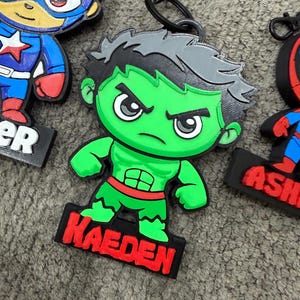 May include: Three colorful rubberized character keychains featuring stylized versions of Captain America, the Hulk, and Spider-Man. Each figure is attached to a black plastic clip and includes a black base with the names Asher and Kaeden.