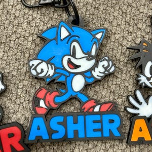 May include: A blue and white cartoon character keychain with the name "ASHER" in blue letters. The keychain has a black clip and is made of a rubbery material. The character is running and has red shoes.