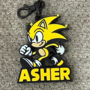 May include: A black and yellow keychain featuring a cartoon character with the name "ASHER" in yellow block letters. The character has yellow spiky hair, black and white shoes, and is in a running pose. The keychain has a black clasp.