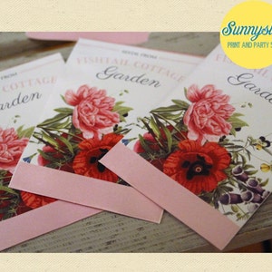 May include: Three pink and white seed packets with floral illustrations and the text "Fishtail Cottage Garden" on the front. The packets are on a wooden surface.
