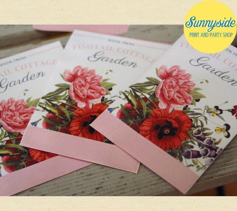 May include: Three pink and white seed packets with floral illustrations and the text "Fishtail Cottage Garden" on the front. The packets are on a wooden surface.