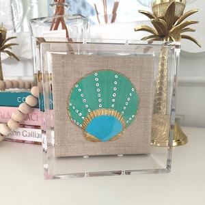 May include: A square clear acrylic display box containing a beige linen-textured square panel. Centered on the panel is a painted seashell in shades of teal and bright blue, featuring gold-painted ridges and vertical rows of small white dots.
