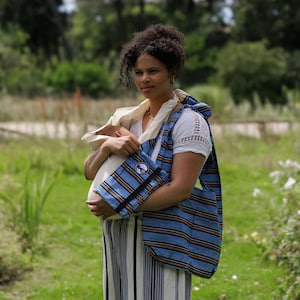 Könnte beinhalten: Eine Frau trägt eine Babytrage mit blau-braunen Streifen und eine passende Tasche. Sie hält ein Baby, das in eine cremefarbene Decke gewickelt ist. Der Hintergrund ist eine grüne Wiese mit Bäumen.