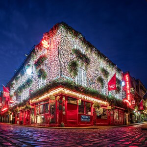 May include: Nighttime exterior shot of a pub in Dublin, Ireland. The building is covered in lights and greenery, with red accents and signage. The street is cobblestone, and the sky is a deep blue.