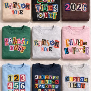 May include: Nine crew neck sweatshirts in various colors, each with unique text designs. The sweatshirts feature phrases like "COOL VIBES ONLY", "2026", and "CUSTOM TEXT", along with numbers and the alphabet. The text is in a colorful, block letter style.
