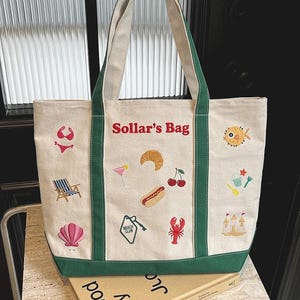 May include: Cream-colored tote bag with green trim and handles, embroidered with beach-themed icons like a bikini, seashell, and cocktail. The text "Sollar's Bag" is embroidered in red. The bag is sitting on top of a book.