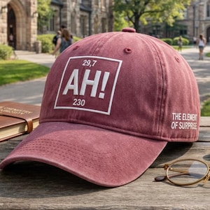 May include: A maroon baseball cap with the text "AH!" in a white box, along with the numbers "29,7" and "230". The phrase "THE ELEMENT OF SURPRISE" is printed on the side. The hat is sitting on a wooden table.