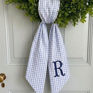 May include: A blue and white gingham wreath sash with a navy blue embroidered letter "R". The sash is tied to a green wreath and hangs on a white door. The sash is made of a soft fabric.