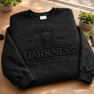 May include: Black sweatshirt with the text "HELLO DARKNESS MY OLD FRIEND" in raised lettering. The design includes a pint of beer with a shamrock and the year 1759. The sweatshirt is on a wooden surface.