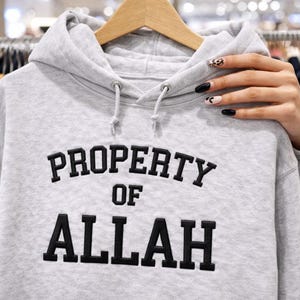May include: A light gray hoodie with the text "PROPERTY OF ALLAH" in black, displayed in a collegiate font. The hoodie has a drawstring hood and is hanging on a wooden hanger.