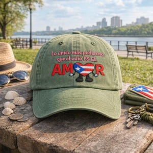 May include: Olive green baseball cap with embroidered text and a heart design. The text reads "Lo único más poderoso que el odio ES EL AMOR" with a heart featuring the Puerto Rican flag. A straw hat and sunglasses are visible in the background.
