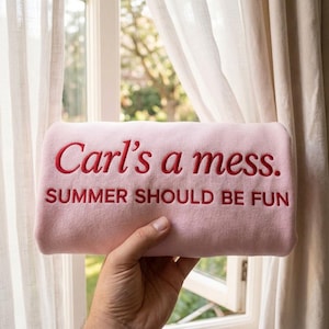 May include: A light pink rectangular towel with the embroidered text "Carl's a mess." and "SUMMER SHOULD BE FUN" in red. The towel is held in front of a window with white curtains.