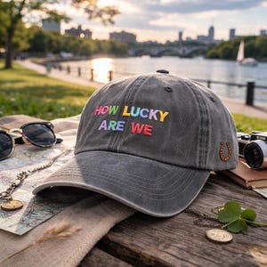 May include: A gray baseball cap with the embroidered text "HOW LUCKY ARE WE" in rainbow colors. The cap features a gold horseshoe emblem and is set on a wooden surface with sunglasses, a map, and a camera.
