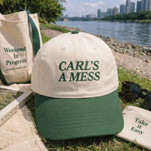 Peut inclure: Casquette de baseball crème et verte avec l'inscription brodée "CARL'S A MESS". La casquette fait partie d'un ensemble comprenant un sac fourre-tout avec le texte "Weekend In Progress" et une petite pochette avec "Take it Easy".
