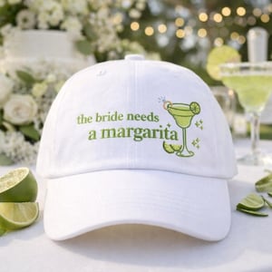 May include: White baseball cap with the text "the bride needs a margarita" in green. The cap features an embroidered margarita glass design with lime slices and sparkles. Limes and a margarita cocktail are in the background.