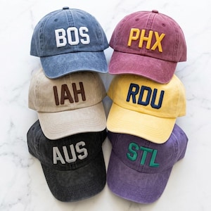 May include: A stack of six baseball caps in various colors, including blue, burgundy, tan, yellow, black, and purple. Each cap features a different three-letter city abbreviation in a contrasting color: BOS, PHX, IAH, RDU, AUS, and STL.